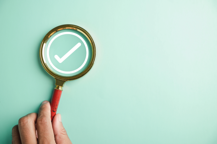 Eine Hand hält eine goldene Lupe über einem weißen, eingekreisten Haken-Symbol (Checkmark) auf helltürkisem Hintergrund, was Kontrolle, Verifizierung oder Erfolg symbolisiert.